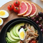 A vibrant, healthy salad bowl with avocado, eggs, tomatoes, grapefruit, and cherries on a wooden board.
